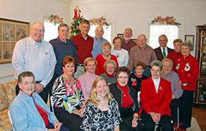 A group of adults smiling and posing together in a living room decorated for the holidays. A Christmas tree is in the background. The group includes both men and women, dressed in casual and festive attire.