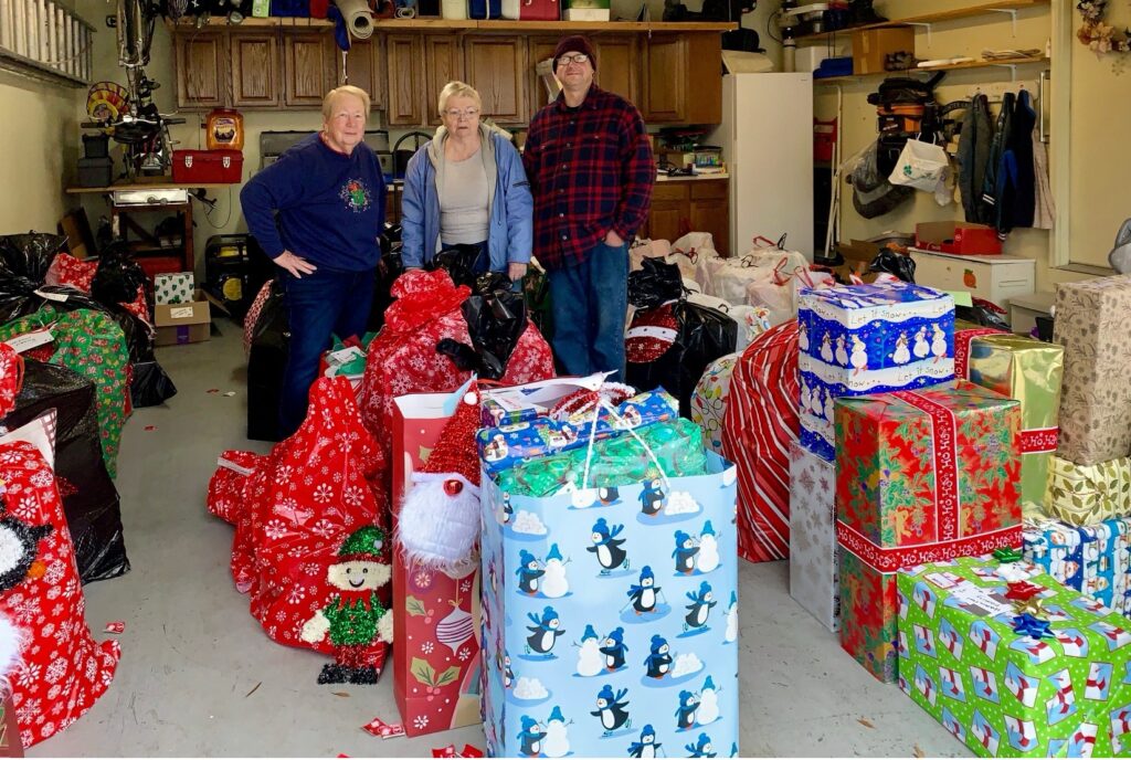 Three people stand in a garage filled with large bags and wrapped presents, including festive gift bags and boxes with holiday patterns, surrounded by shelves and tools along the walls.