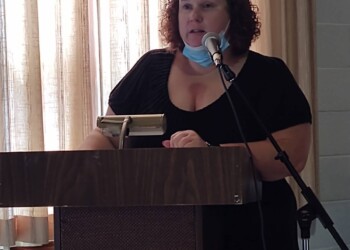 A woman with curly hair stands at a podium, speaking into a microphone. She wears a black top and has a blue face mask pulled down below her chin. Light filters through curtains behind her.