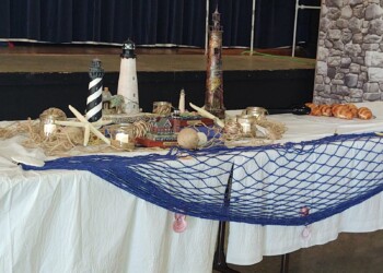 A table decorated with miniature lighthouses, seashells, starfish, netting, and candles, with some pastries on the side. The backdrop includes a stage and a wall with a stone pattern.