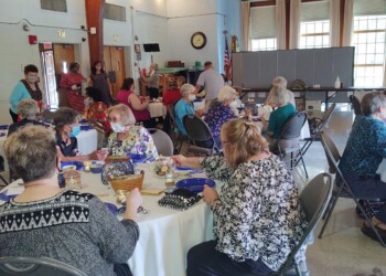 A group of people sits around round tables in a hall with high wooden ceilings, eating and talking. Some people are serving food from a buffet table in the back. The atmosphere appears friendly and communal.