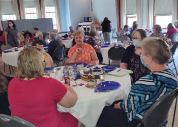 A group of women sit around round tables covered with white tablecloths, eating and talking in a large room with high wooden ceilings and big windows. Some attendees are wearing face masks.