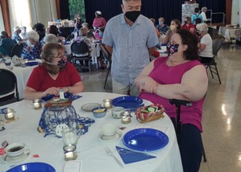 A group of people wearing masks sit around tables with blue plates and teacups at a social gathering in a hall. A man stands talking to two seated women at a decorated table. Candles and snacks are on the table.