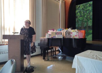 A woman stands at a podium with a microphone in a sunlit room. Behind her is a piano covered with colorful gift bags and a painted backdrop of green trees. Tables and chairs are visible in the foreground.