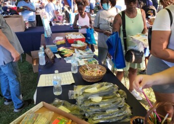 People gather around outdoor tables under tents at a community event. Tables display school supplies like pencils, erasers, and bottled water. Attendees are browsing and selecting items. Trees and more booths are visible in the background.
