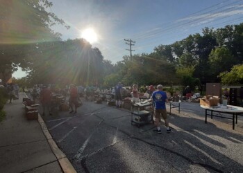 People browse tables with various items for sale at an outdoor market on a sunny day. The scene is set in a parking lot with trees and power lines in the background. Sunlight creates a lens flare effect.