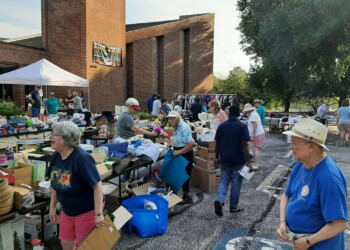 A busy outdoor yard sale takes place in a parking lot beside a brick building, with tables full of items and many people browsing and talking under a sunny sky.