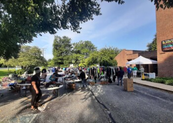 People shop at an outdoor sale in a parking lot beside a brick building. Tables display clothes and various items under sunny skies, with trees and a sign visible in the background.