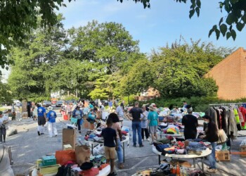 People browse tables with clothes and items at an outdoor yard sale on a sunny day, surrounded by trees and a brick building. Shoppers and vendors interact in a busy, lively atmosphere.
