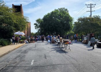 People are browsing and shopping at an outdoor yard sale set up on a closed street lined with various furniture and household items, with trees and a building in the background under a clear sky.