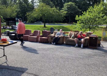 Several pieces of furniture, including sofas and chairs, are displayed outside on pavement. Three people sit on a couch, while another person walks by. Trees and a road with cars appear in the background.