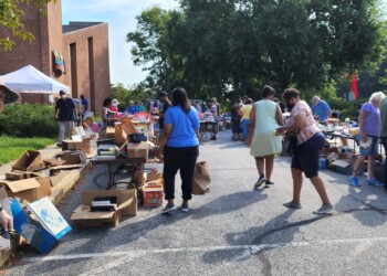 People browse tables of items at an outdoor yard sale on a sunny day. Cardboard boxes are scattered around, and various goods are displayed. Trees and a brick building are in the background.