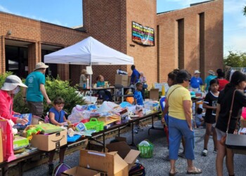 People browse tables filled with various items at an outdoor yard sale in front of a brick building. Some are shopping, while others manage the tables under a white canopy on a sunny day.