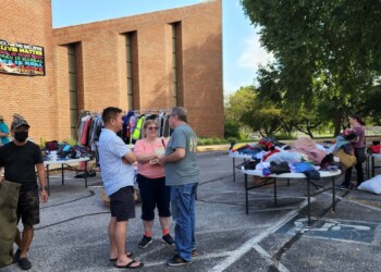 People browse and talk at an outdoor clothing giveaway event in a parking lot beside a brick building, with tables of clothes and trees providing shade. A colorful sign is visible on the building in the background.