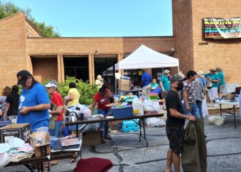 A group of people shop and interact at an outdoor yard sale in front of a brick building with tables displaying various items. A colorful banner hangs on the building, and a white canopy tent provides shade.