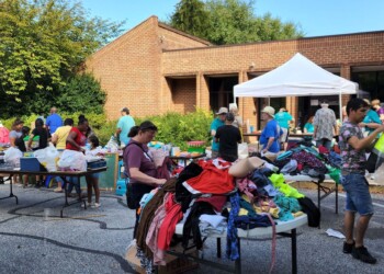 People browse tables filled with clothes and household items at an outdoor community yard sale held in front of a brick building on a sunny day. A white canopy tent is set up and trees are visible in the background.