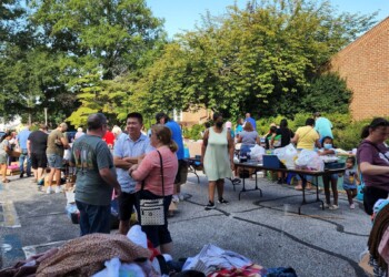People gather outdoors at a community event, sorting and collecting clothes and items from tables and piles in a parking lot surrounded by trees and brick buildings on a sunny day.