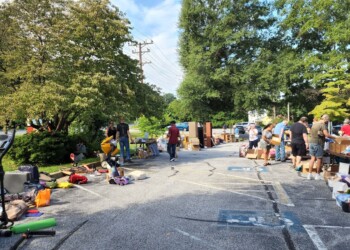 People browse and sell various items at an outdoor yard sale on a sunny day. Tables and goods line both sides of a paved parking lot, with scattered items on the ground and trees in the background.