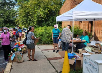 People gather outside a brick building at a community event. Some are browsing tables with goods, while others talk near a white tent where volunteers sit behind a table. Trees and greenery are visible in the background.
