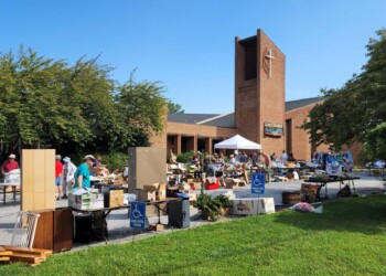 Outdoor yard sale in front of a brick church with a cross on the tower. Tables display various items for sale, and people browse under a clear blue sky. Trees and grass frame the scene.