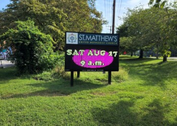 A digital sign outside St. Matthew’s United Methodist Church displays “SAT AUG 17 9 A.M.” in bright colors, surrounded by grass, trees, and a small outdoor market on the left.