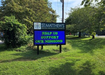 A digital sign outside St. Matthew’s United Methodist Church reads, “HELP US SUPPORT OUR MISSIONS,” surrounded by green grass and trees on a sunny day.