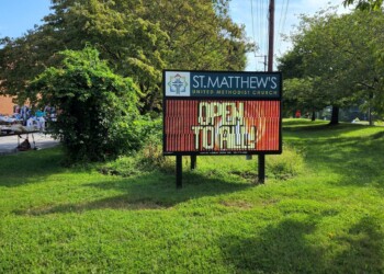 A digital sign on a grassy lawn outside St. Matthews United Methodist Church reads Open to All. People and tables are visible in the background near the church building.
