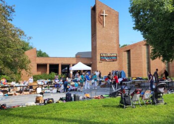 People gather outside a modern brick church with a tall cross, attending a community sale or market with tables of items, strollers, and a banner welcoming all on the building. Trees and grass frame the scene.