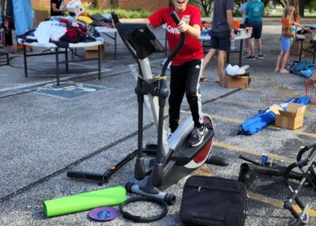 A child in a red hoodie stands on an elliptical machine at an outdoor yard sale, surrounded by various items on the ground and other people browsing in the background. Trees and a brick building are visible.