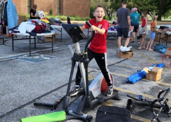 A child in a red shirt smiles while using an elliptical machine at an outdoor yard sale, with scattered clothes and equipment nearby and people browsing tables in the background.