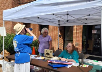 Three people are at an outdoor table under a white canopy. One person in a hat drinks from a bottle, while two others sit and stand behind the table, which has papers, clipboards, and supplies on it, outside a brick building.