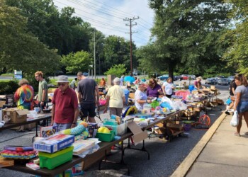 People browse and shop at an outdoor yard sale, with tables full of assorted items like toys, games, and household goods. Trees, parked cars, and a sunny sky are visible in the background.