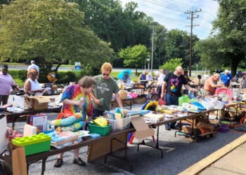 People browse tables filled with toys, books, and various items at an outdoor yard sale on a sunny day, with trees and a road in the background.