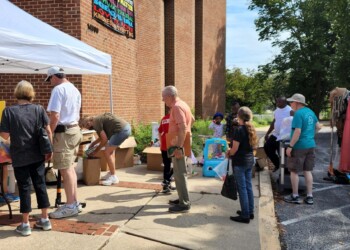 People browse and shop at an outdoor sale with tables, boxes, racks of clothes, and various items on display near a brick building and under a white canopy tent. Trees and sunlight are visible in the background.
