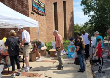 People browse and shop at an outdoor yard sale next to a brick building, with tables, racks of clothes, and various items for sale under a white canopy tent on a sunny day.