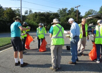 A group of people wearing safety vests and hats gather in a parking lot, holding orange trash bags and litter pickers, preparing for a community clean-up on a sunny day.
