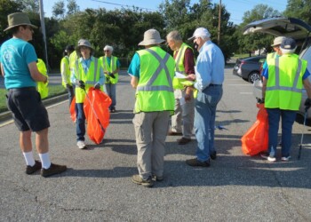 A group of adults wearing yellow safety vests and hats gather in a parking lot holding orange bags, preparing for a community cleanup event on a sunny day.