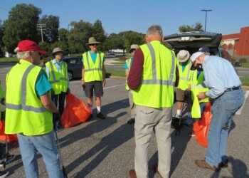 A group of people wearing yellow safety vests gathers in a parking lot with orange trash bags, preparing for a community clean-up on a sunny day.