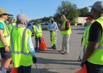 A group of people wearing yellow safety vests gathers in a parking lot, holding orange trash bags and litter pickers, preparing for a cleanup event on a sunny day.