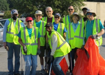 A group of people wearing yellow safety vests and hats stand together in a parking lot, smiling and holding orange trash bags and litter pickers, ready for a cleanup effort.