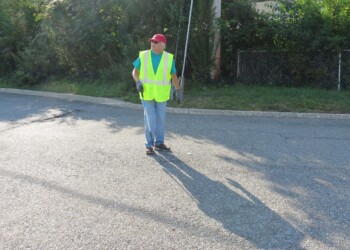 A person wearing a neon yellow safety vest, red cap, gloves, and blue jeans stands on a sunlit paved street, holding a litter picker with trash in the background and greenery behind them.