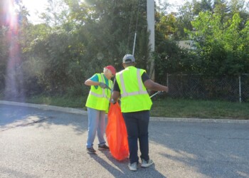 Two people wearing yellow safety vests and gloves pick up litter with grabbers and place it in an orange bag on the side of a road, with trees and a fence in the background.