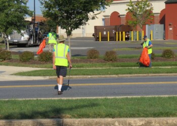 Three people in yellow safety vests pick up litter along a roadside and grassy area, using grabber tools and holding orange trash bags. A building and a truck are visible in the background.