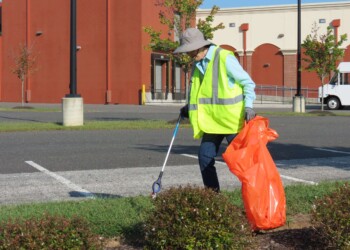 A person wearing a hat, mask, and bright yellow safety vest picks up litter in a parking lot using a grabber tool, holding a large orange trash bag. Bushes and a red building are in the background.