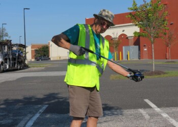 An older man wearing a safety vest, gloves, and a hat uses a grabber tool to pick up litter in a parking lot on a sunny day.
