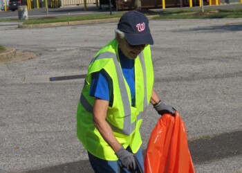 An older person wearing a neon yellow safety vest and a navy cap picks up litter with an orange trash bag in a parking lot.