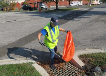 A person wearing a neon safety vest and cap picks up litter with a grabber tool near a storm drain, holding a large orange trash bag in a parking lot area on a sunny day.