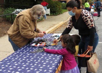 Two adults and two children do crafts at a table covered with a blue cloth outdoors. The children are coloring, while the adults assist. One woman wears a mask, and people are seen in the background.