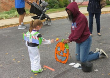 A young child in a Buzz Lightyear costume tosses a beanbag at a Halloween pumpkin target, while an adult in a hoodie kneels nearby on a street. A stroller and other people are in the background.