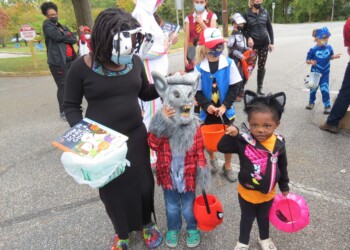 Children in Halloween costumes, including a wolf, a cat, and other characters, stand with adults in a parking lot. They are holding orange buckets and books, ready for trick-or-treating. Trees and parked cars are visible in the background.